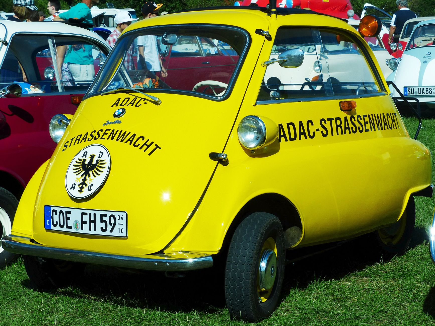 Bright yellow Isetta microcar showcased at an outdoor car exhibition, surrounded by vintage vehicles.