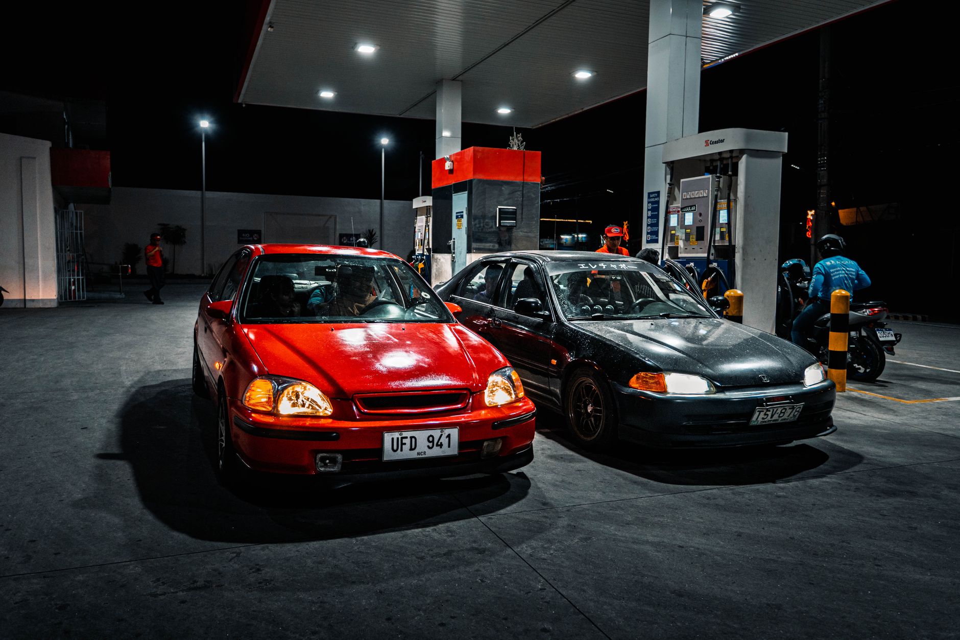 Two cars parked at a gas station at night, illuminated by overhead lights.