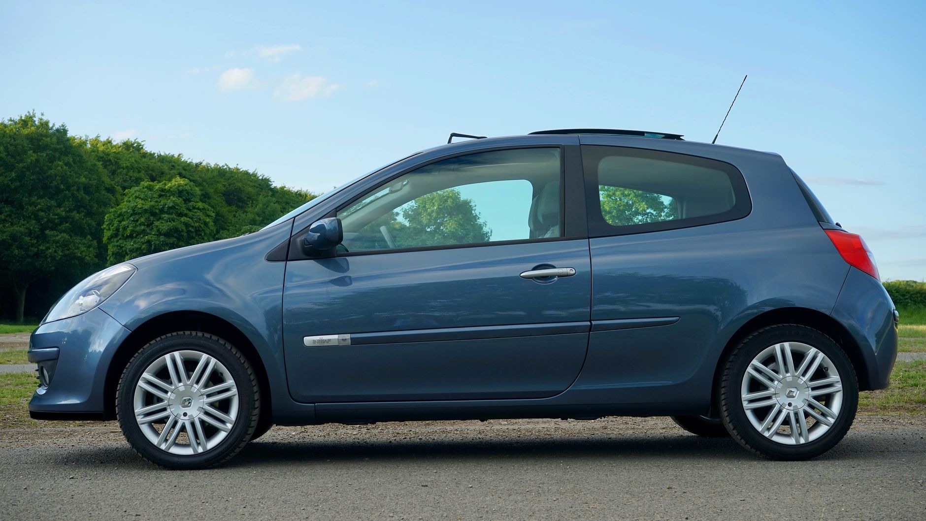 Blue French hatchback car on a country road with a lush green background and clear sky.