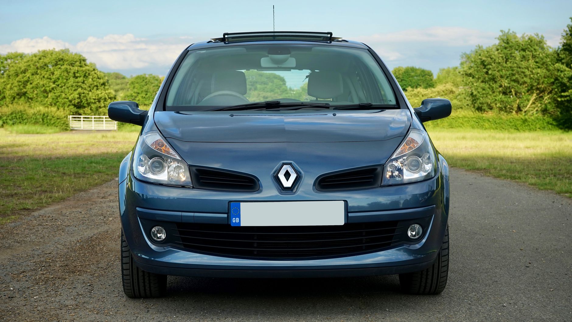 Blue Renault Clio parked on a serene country road, surrounded by lush greenery under a sunny sky.