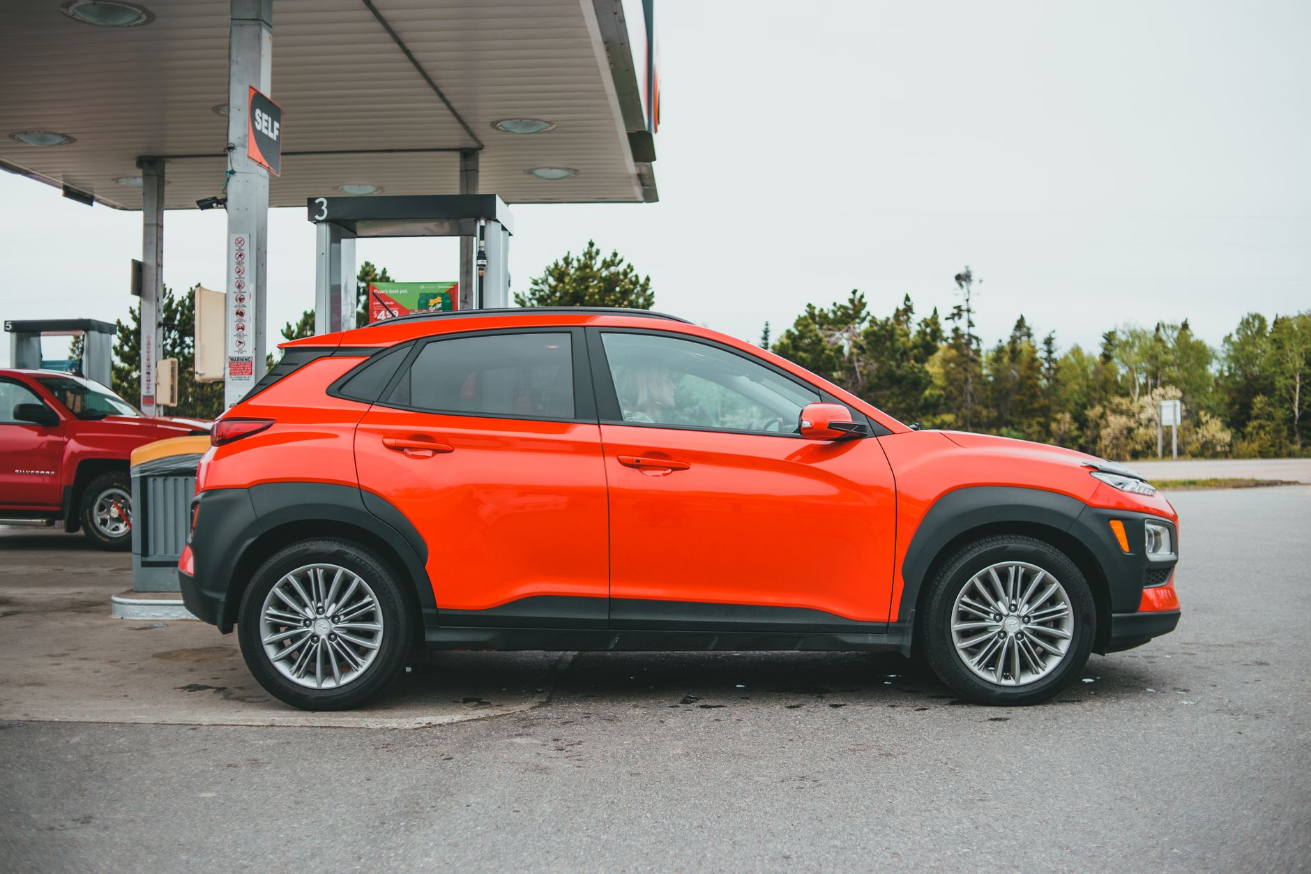 Bright orange SUV parked at a gasoline station with a red truck in the background.