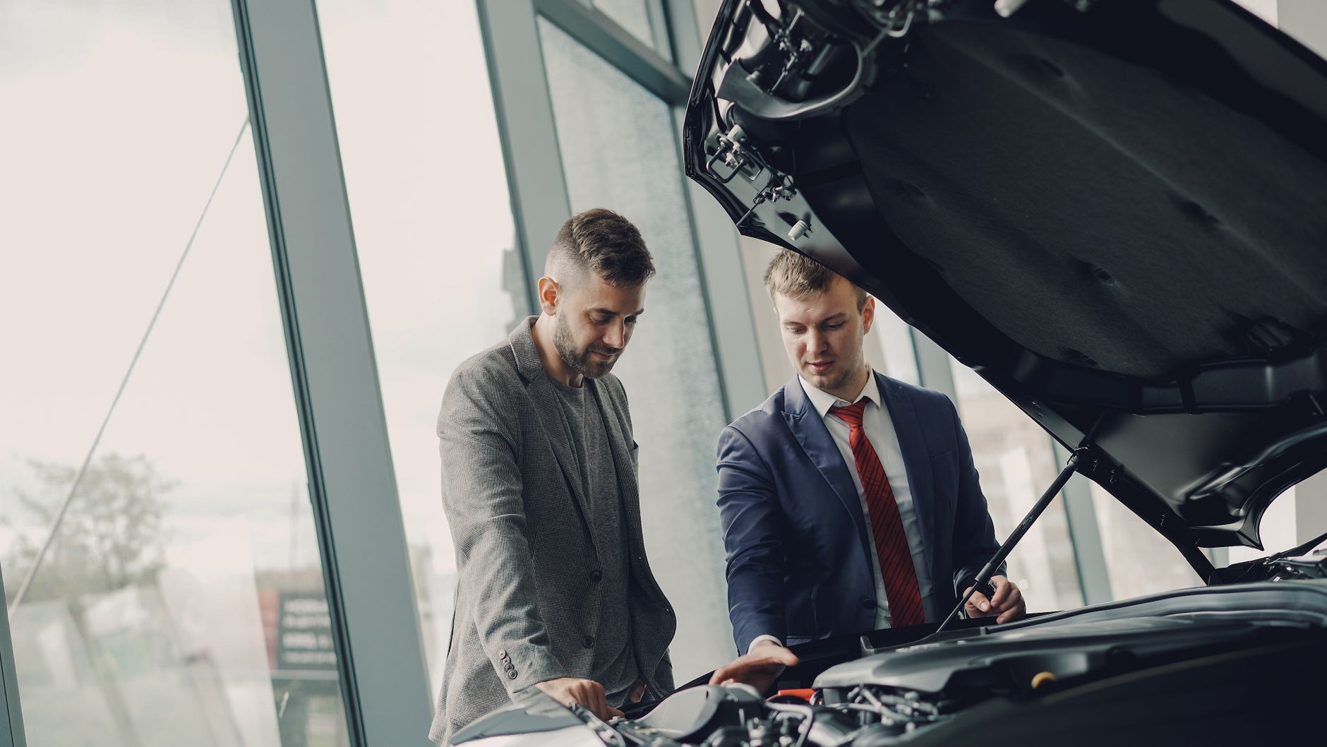 Two men inspecting a car's engine hood in a dealership showroom.