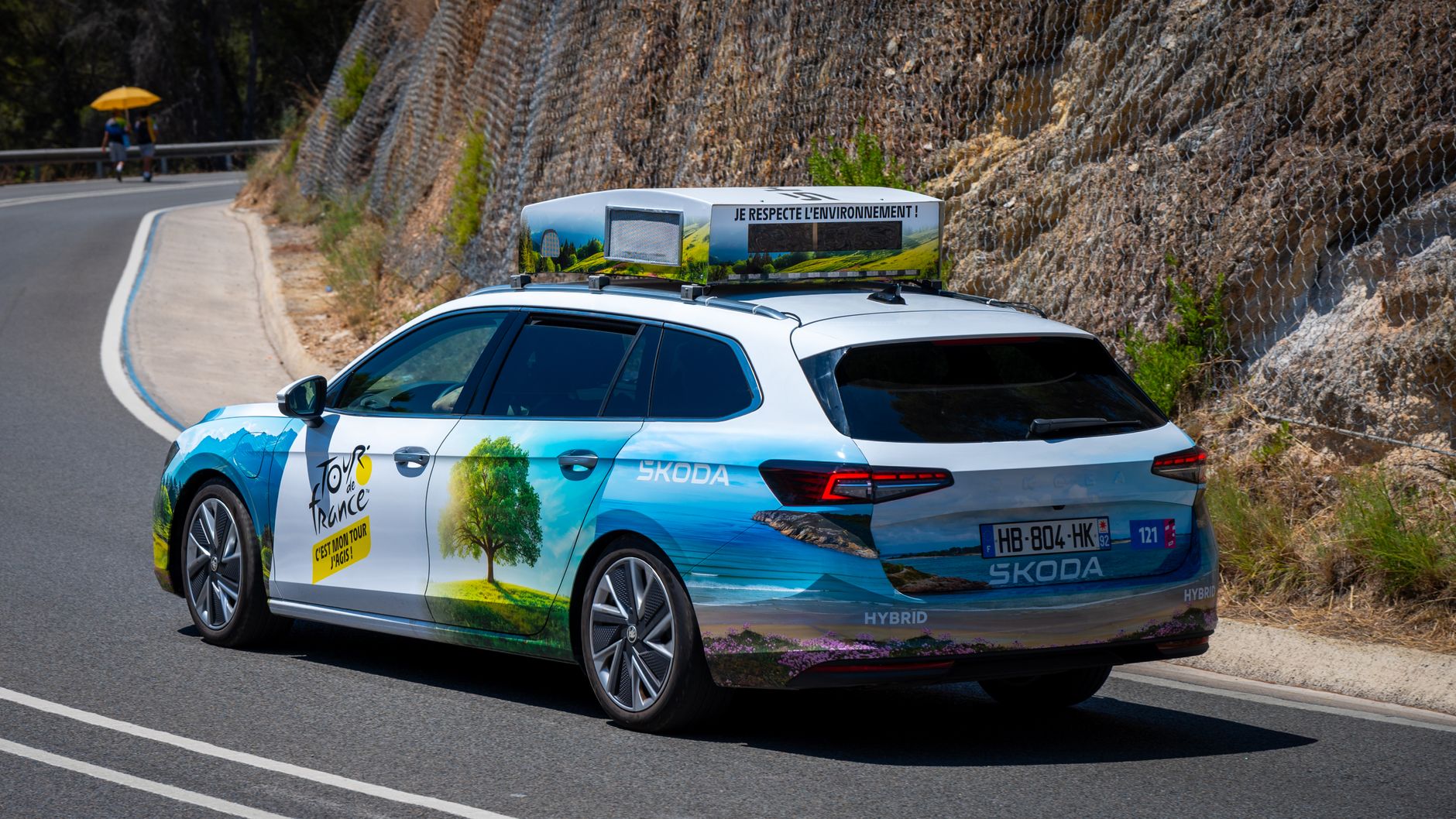 A Skoda hybrid car with Tour de France branding navigates a scenic mountain road.