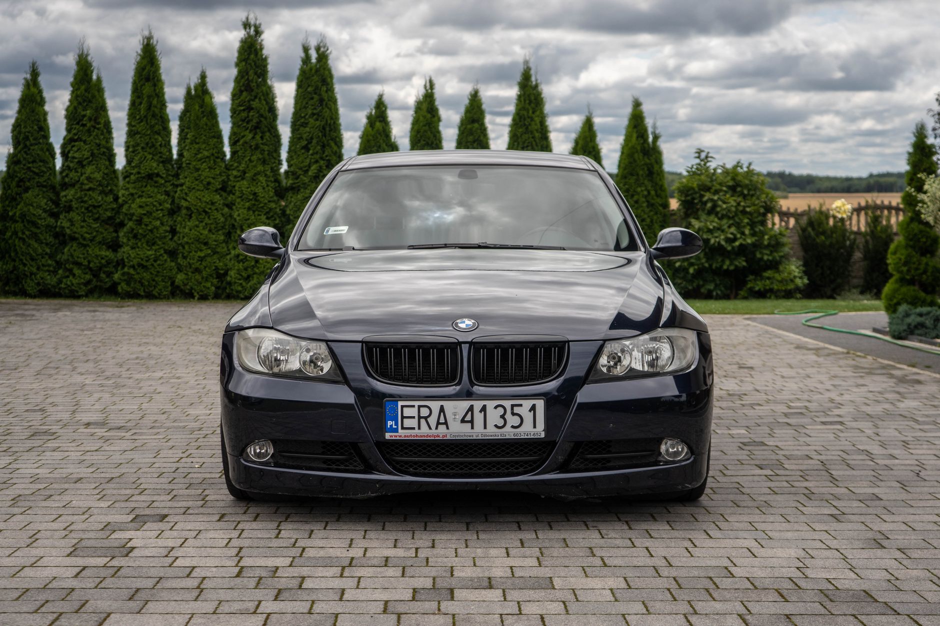 Front view of a BMW 3 Series E90 sedan parked on a paved driveway outdoors.