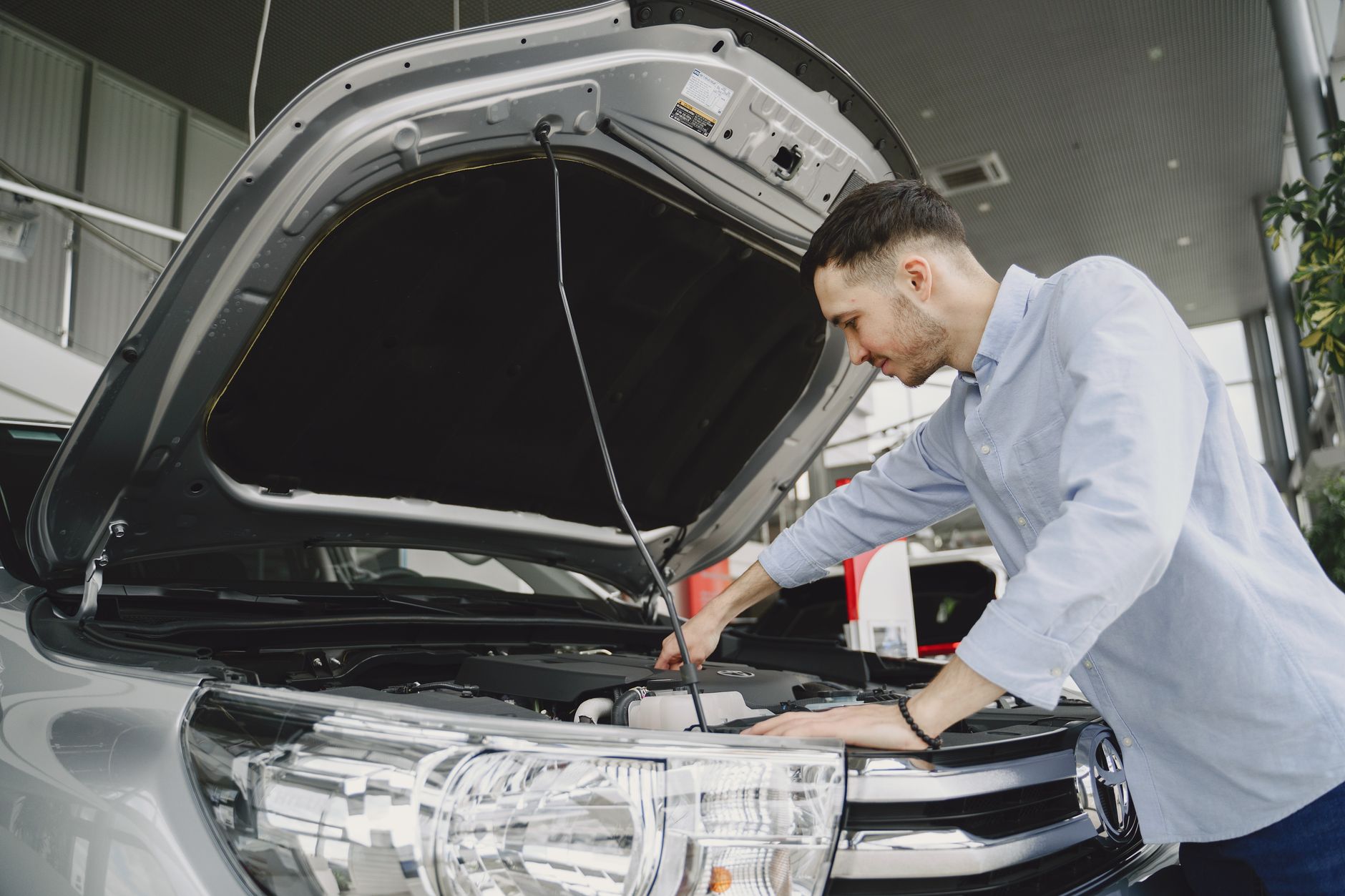A man in a blue shirt examines a car's engine in a modern vehicle showroom.