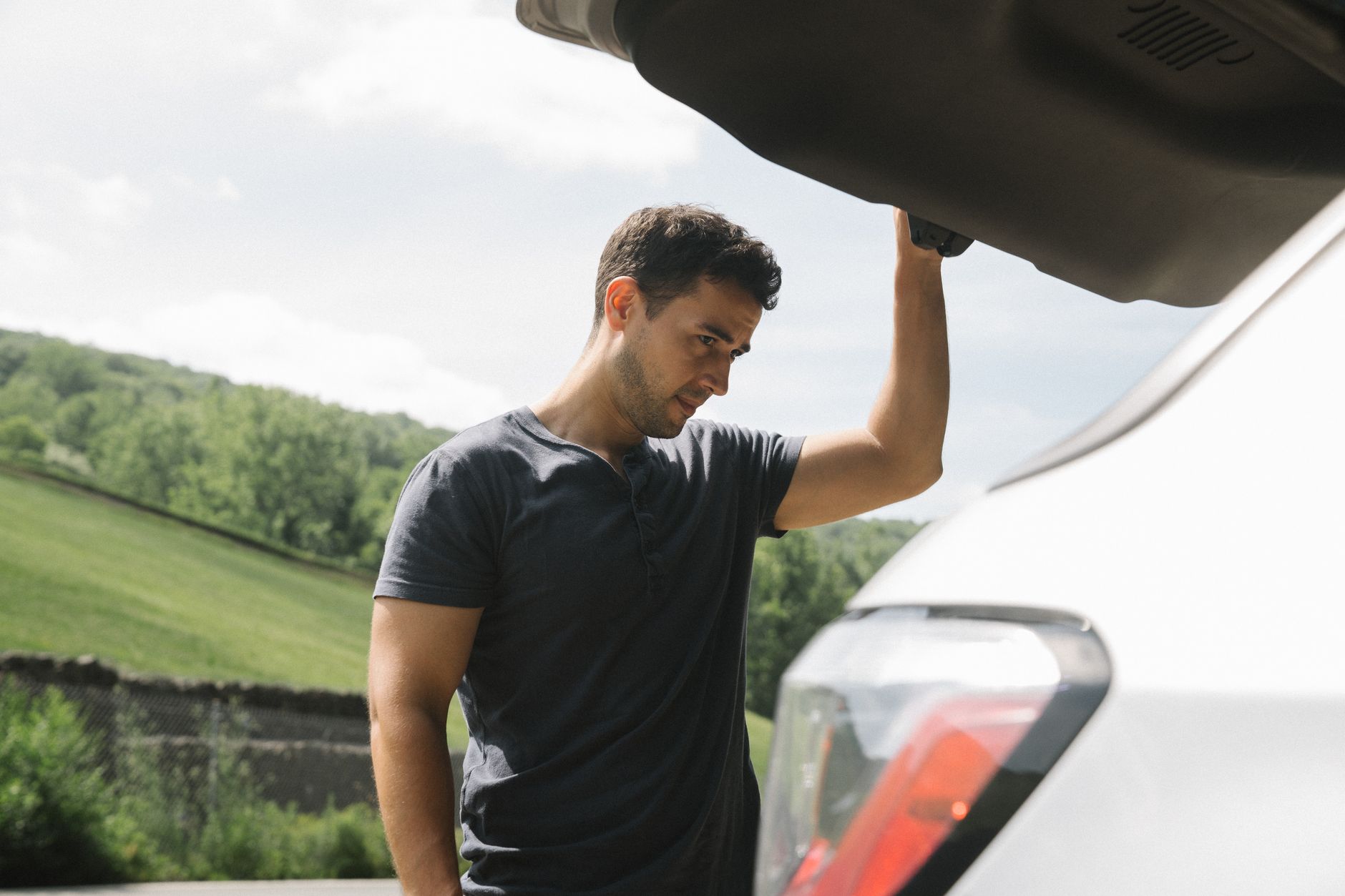 A man examining the open trunk of a car under clear skies with greenery in the background.