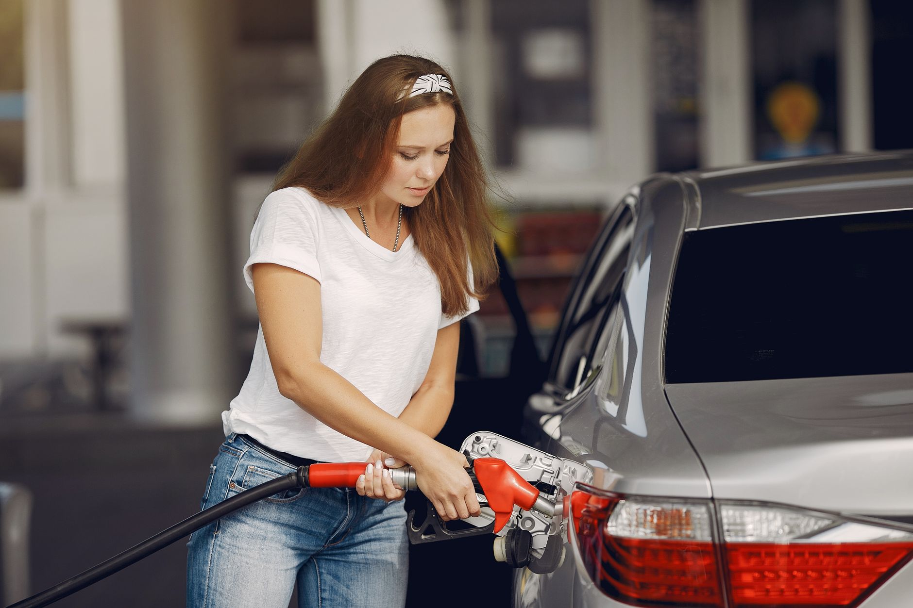 Attentive female driver in casual outfit and headband filling up modern automobile with automotive fuel gun on petrol station while looking 