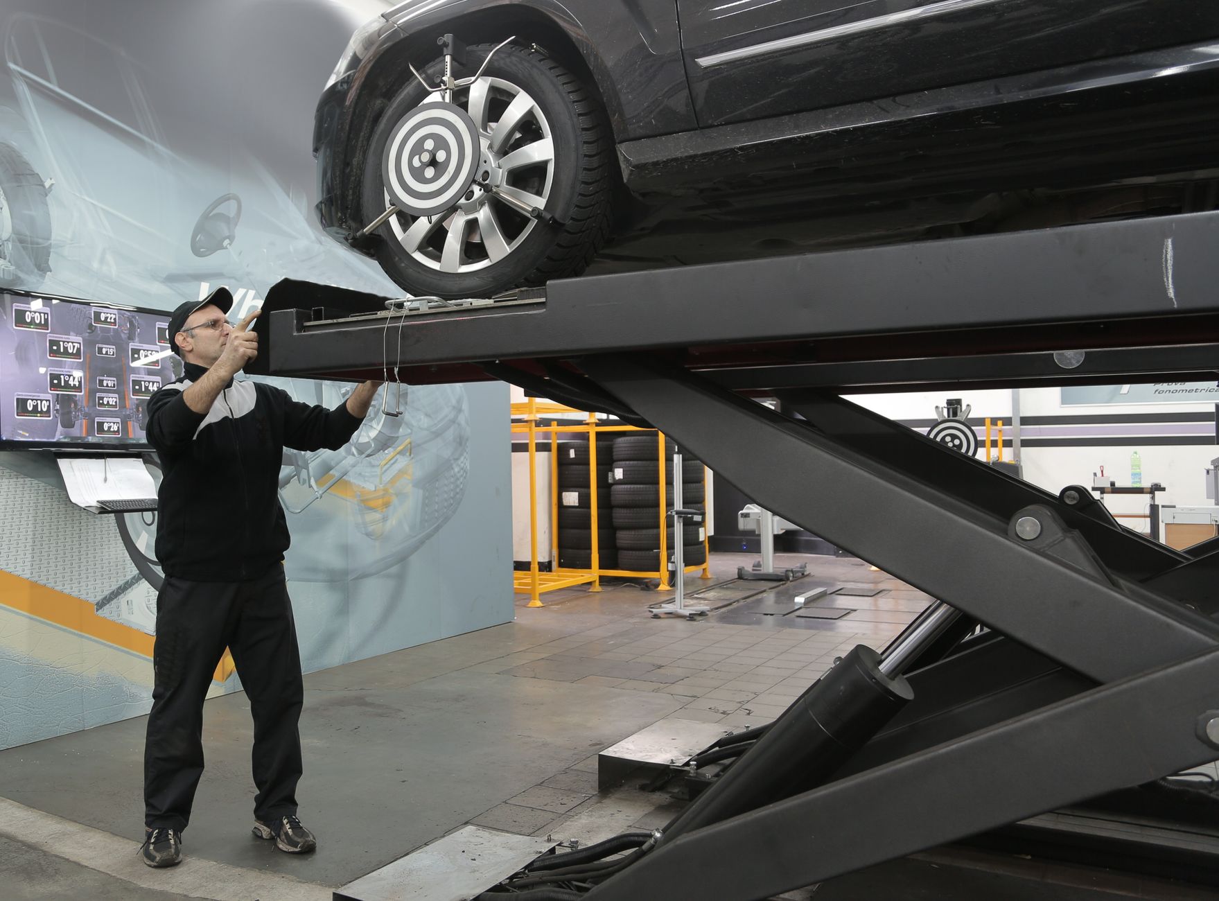 Serious professional adult mechanic in casual uniform examining car wheels while standing near lift in modern car service garage